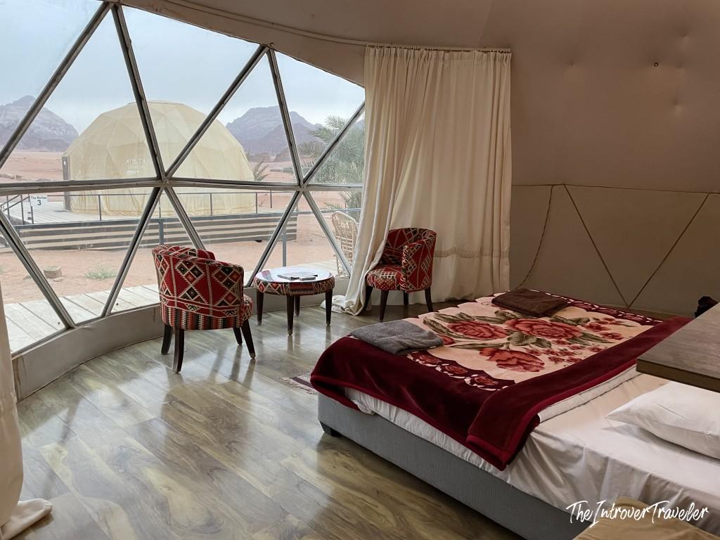 Interior of a Wadi Rum glamping dome, with a double bed, kilim chairs, and desert views through the geodesic window