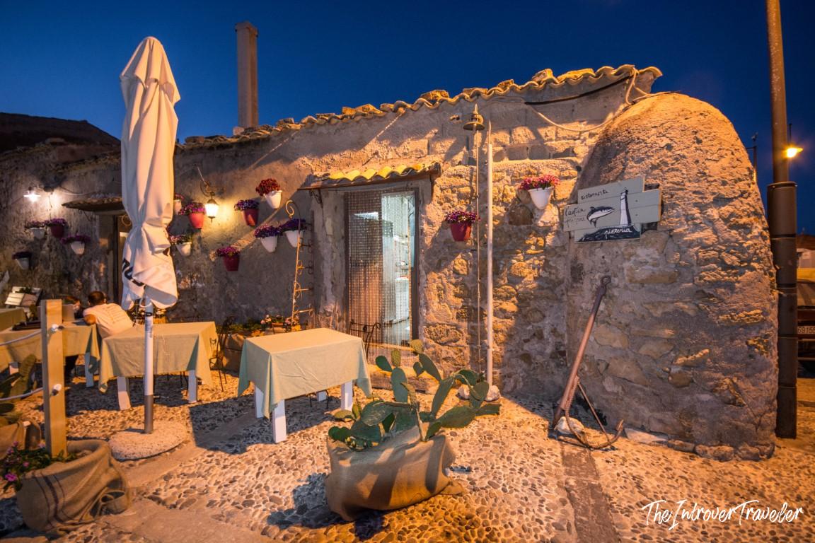 Rustic stone seafood restaurant in Marzamemi Sicily, lit at dusk with flower pots and outdoor tables