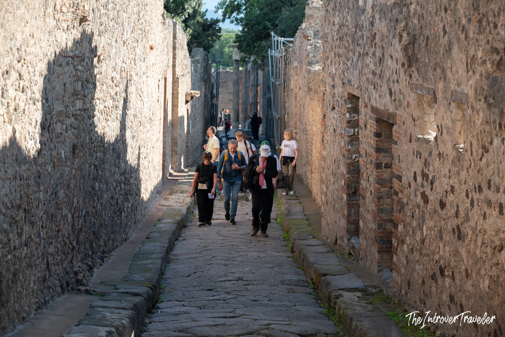 Tourists walking along a narrow ancient Roman street in Pompeii, flanked by stone walls