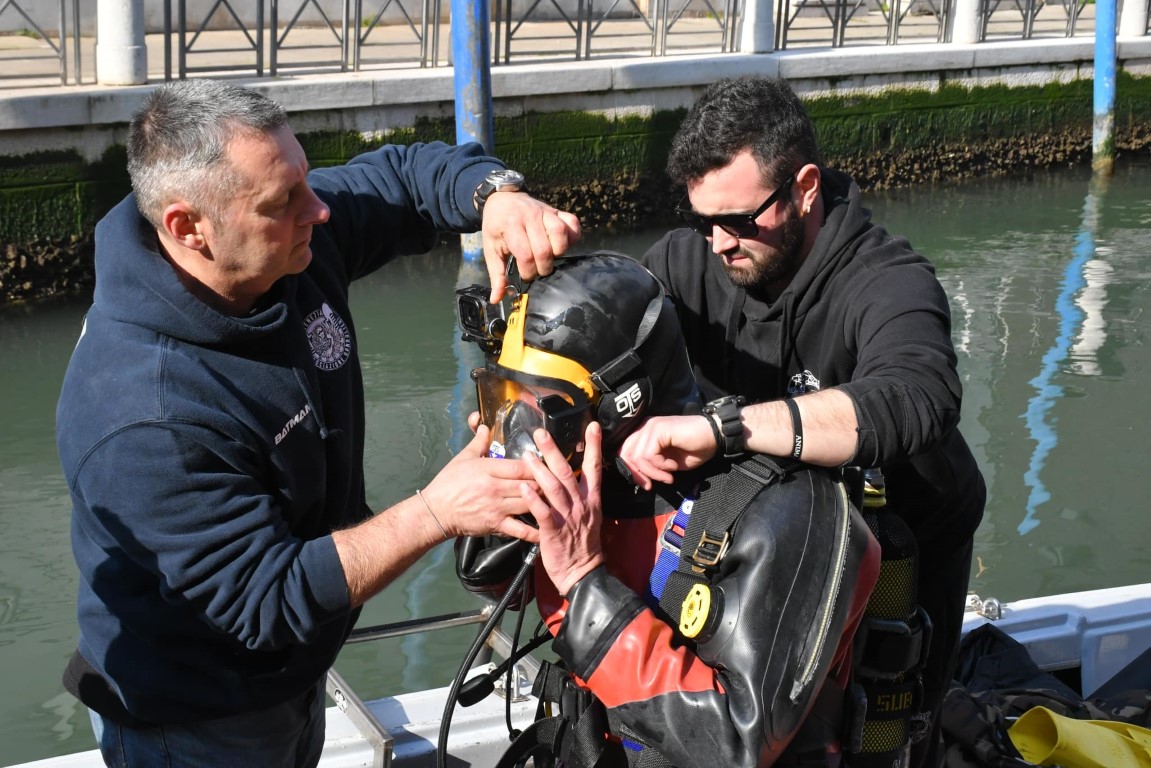 Two Venice canal divers adjusting an OTS diving helmet on a boat in a Venetian waterway