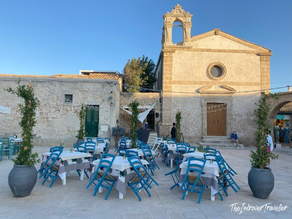 Outdoor restaurant tables with blue chairs in Marzamemi's main piazza, Sicily, beside a Baroque church