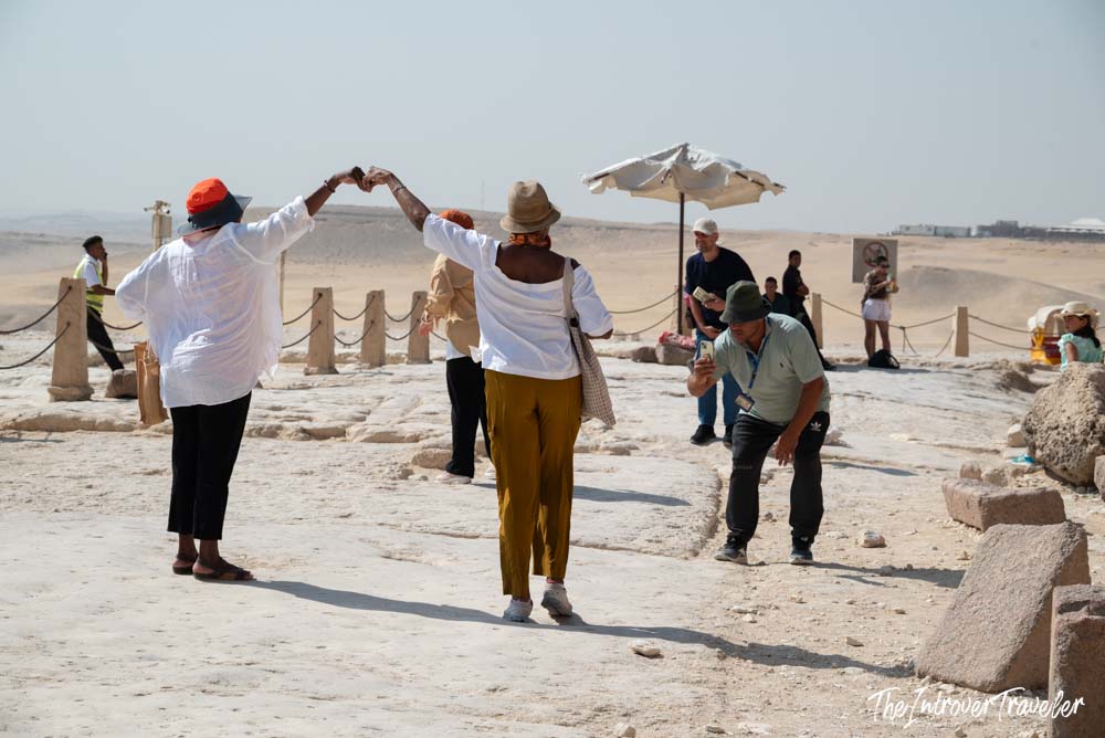 Tourists posing for photos at the Giza plateau, with desert and ancient stone ruins stretching behind them