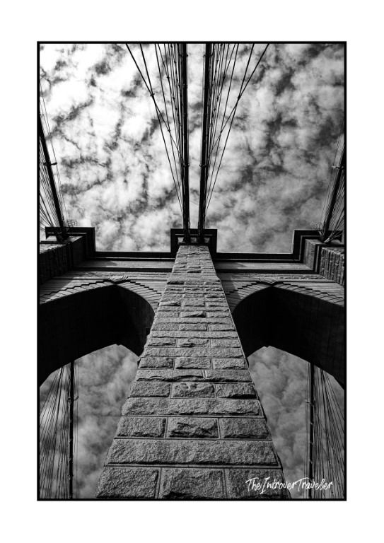Looking straight up at a Brooklyn Bridge tower, cables fanning skyward in black and white