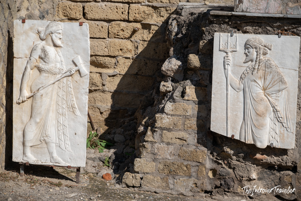 Two ancient Roman marble relief panels displayed against a ruined stone wall at Herculaneum excavations