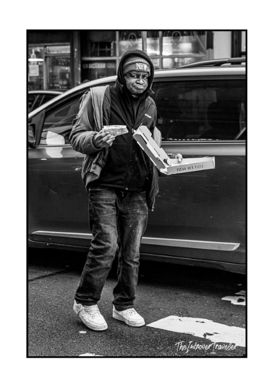 Black and white street photo of a man eating pizza from an open box on a New York City street