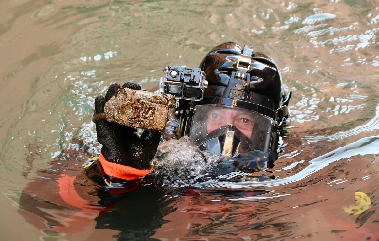 Diver surfacing in a Venice canal, holding a corroded artifact recovered from the murky water below