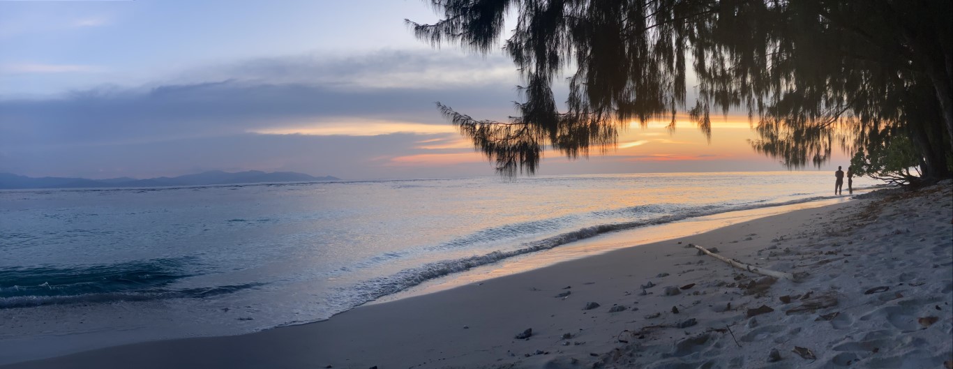 Sunset over a white sand beach in Raja Ampat, with casuarina trees and two silhouetted figures at the shoreline