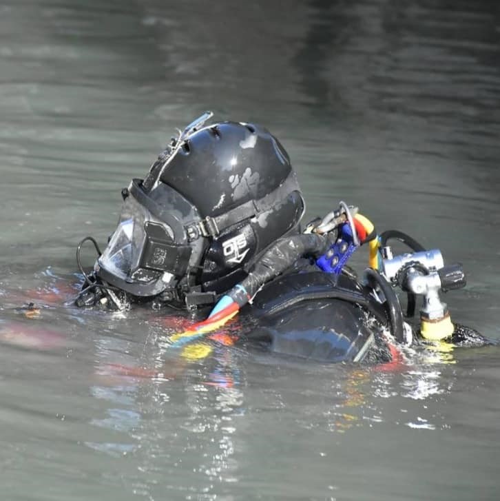 Venice canal diver in full black wetsuit and helmet surfacing in murky gondola waterway