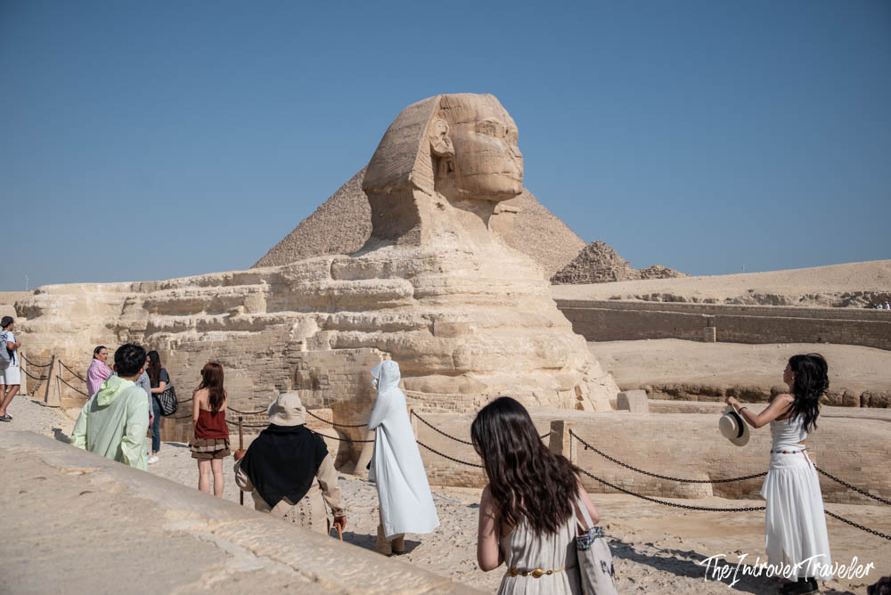 Tourists walking along the viewing path beside the Great Sphinx of Giza, with a pyramid behind