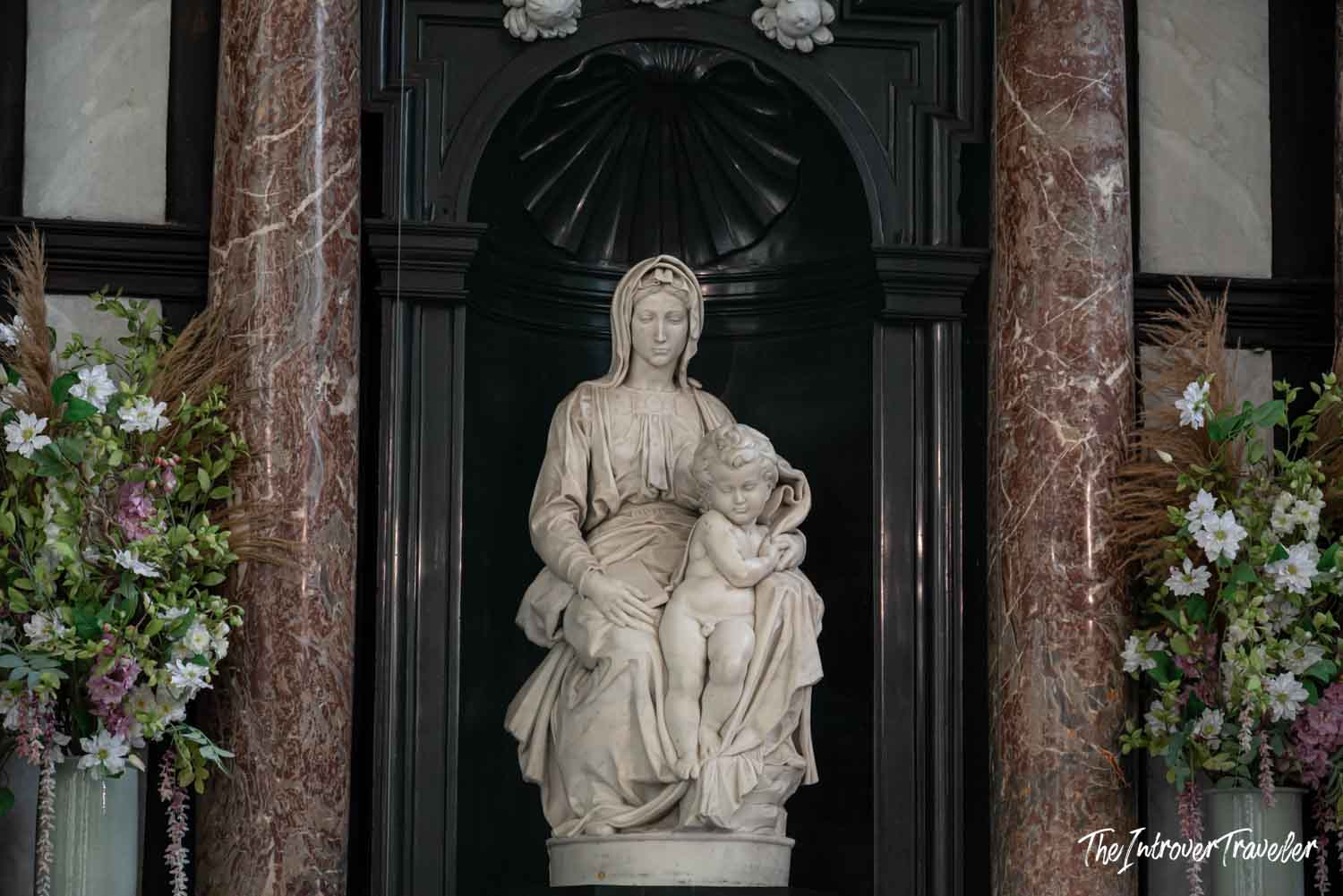 Michelangelo's Madonna of Bruges marble sculpture in a dark niche flanked by floral arrangements