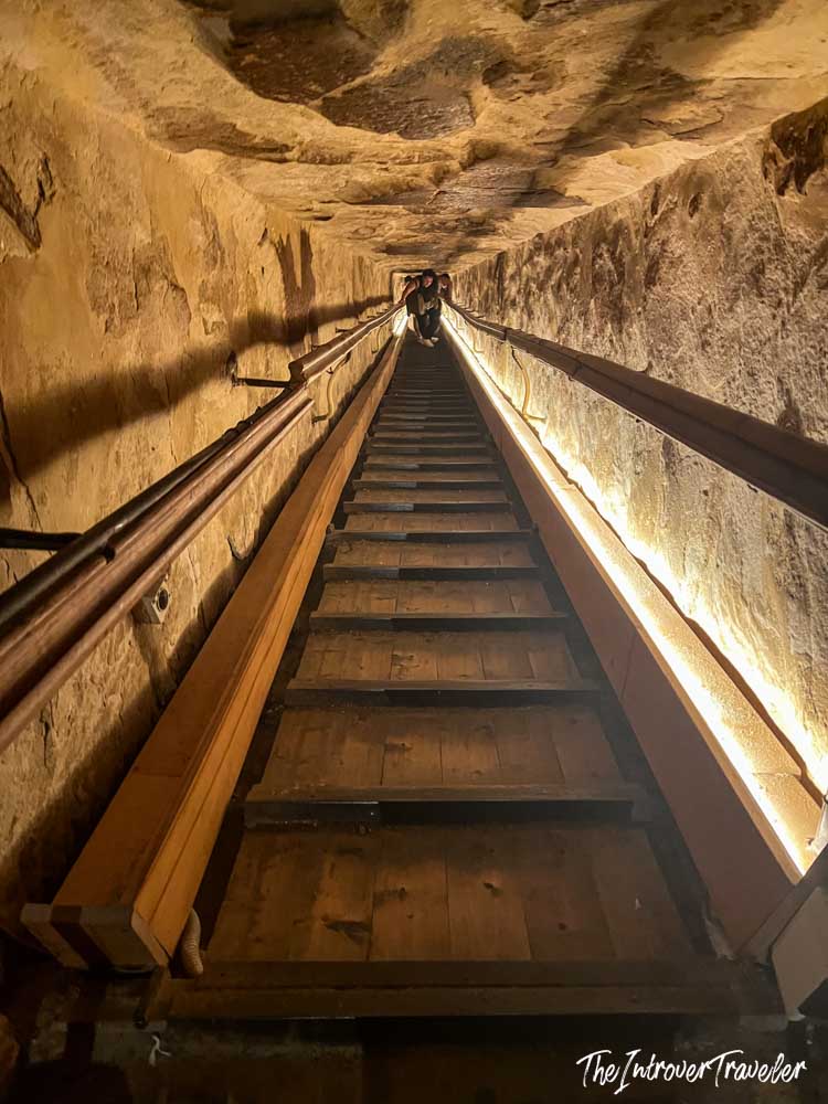 Steep wooden staircase inside a Giza pyramid interior passage, lit by strip lights along carved stone walls