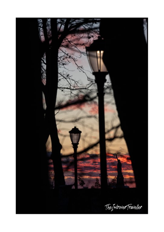Silhouetted lamp posts and bare trees frame a fiery NYC sunset with a distant statue