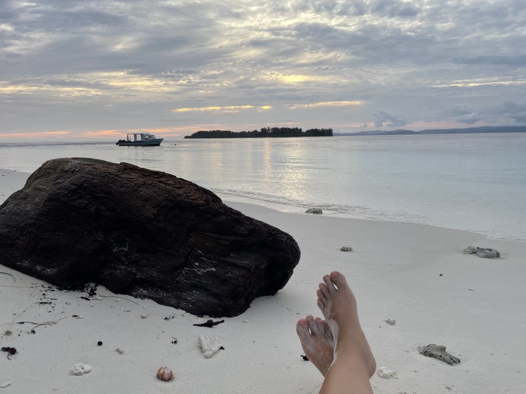 Bare feet resting on white sand beach in Raja Ampat, with small islands and a dive boat at dusk
