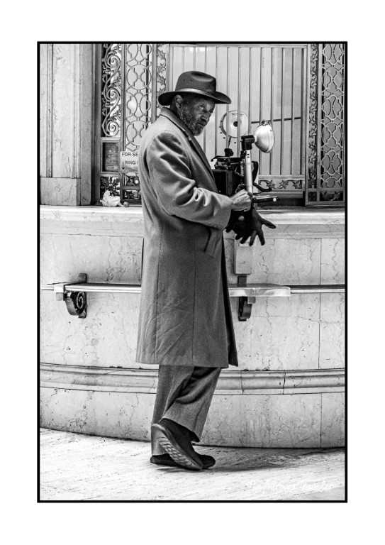 Black and white street photo of a man in coat and fedora at a NYC payphone, December 2024
