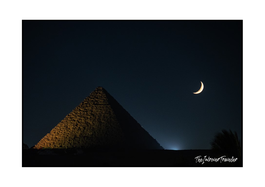 Great Pyramid of Giza at night, its limestone blocks lit against a deep blue sky with a crescent moon above