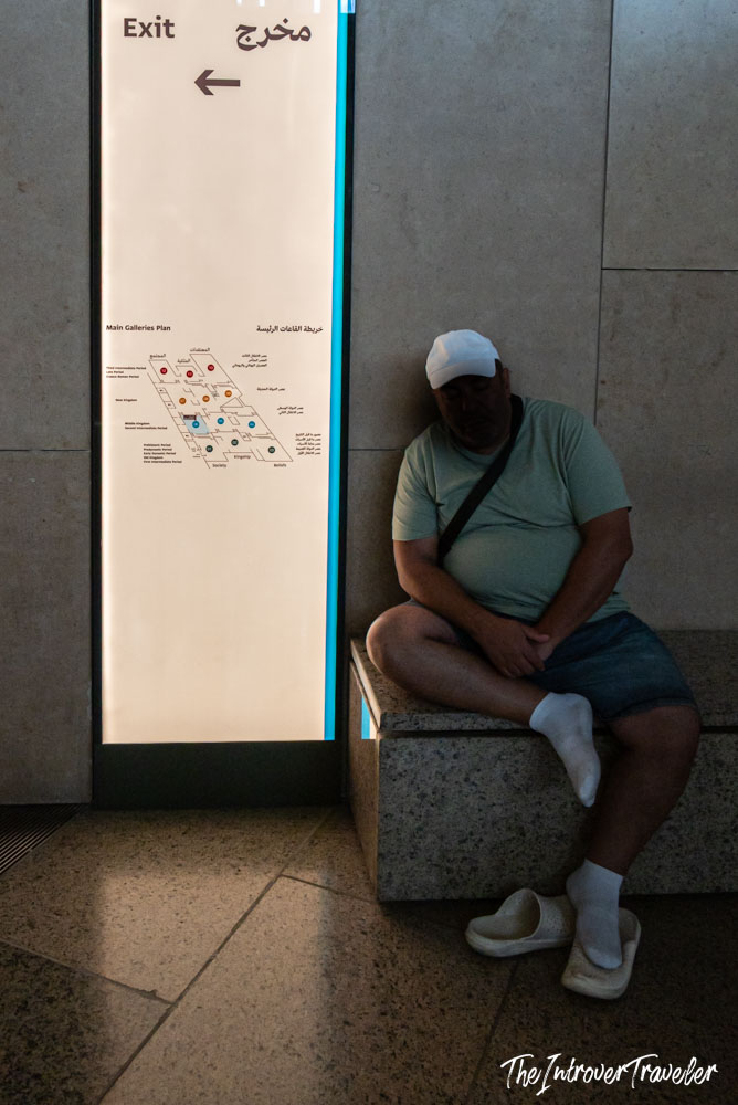 Tired visitor napping beside a bilingual exit sign and gallery map at the Grand Egyptian Museum