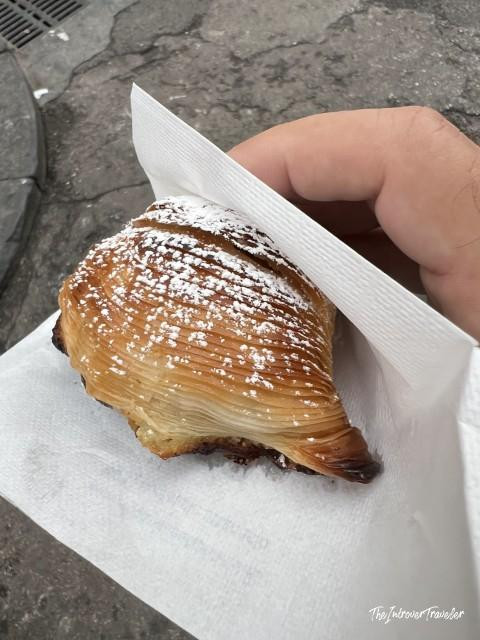 Flaky Neapolitan sfogliatella dusted with powdered sugar, held in a paper napkin on a Naples street