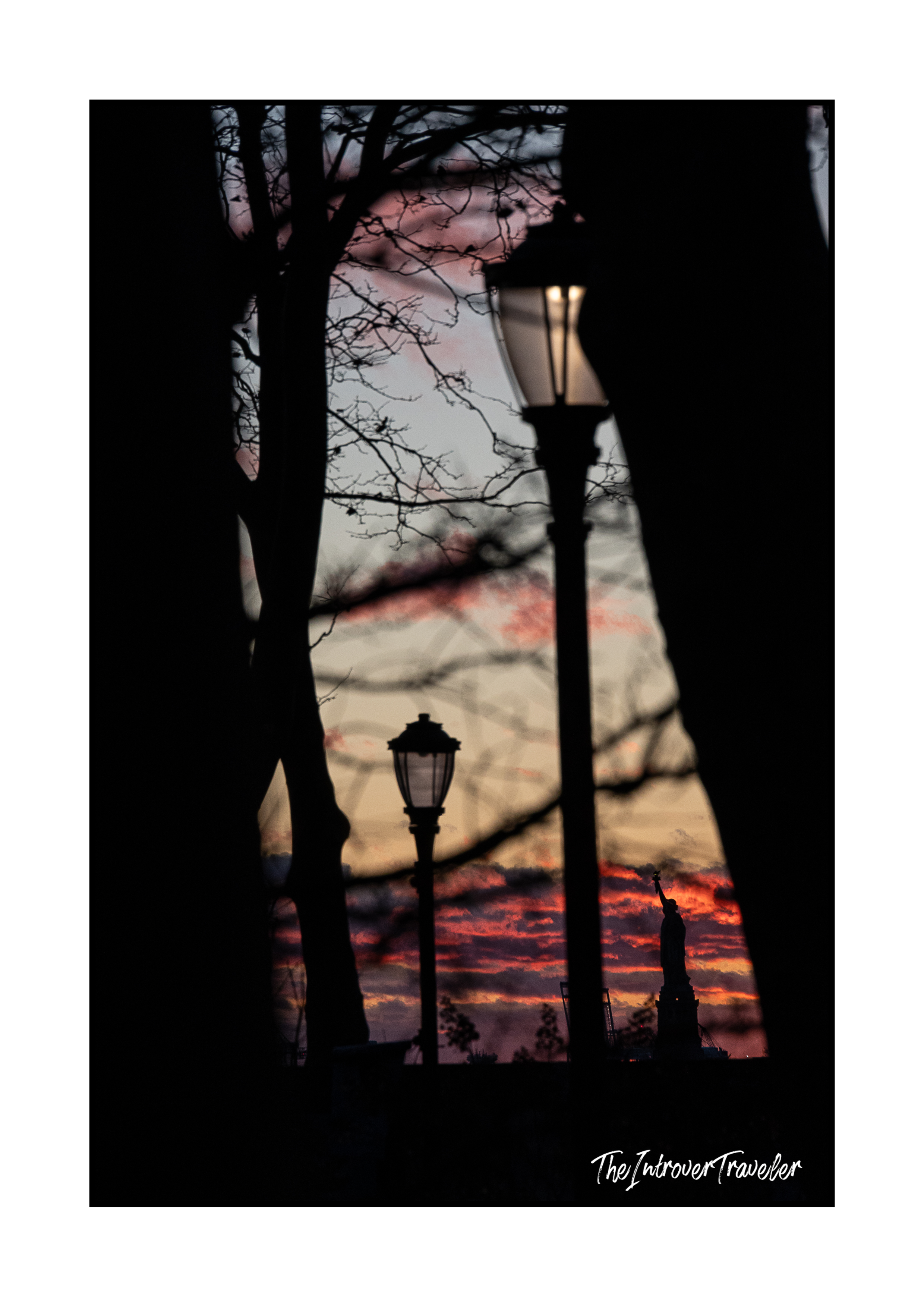 Statue of Liberty silhouette at fiery sunset, framed by bare trees and vintage lampposts in New York