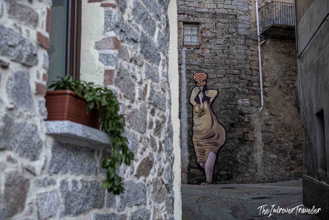 Street art mural of a robed woman with a checkered vessel, painted on a stone wall in Orgosolo, Sardinia