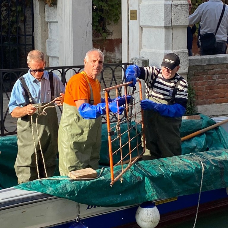 Three Venice canal workers in waders handling a metal grate on a working boat along a Venetian canal