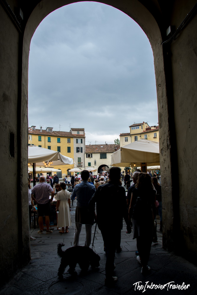 Arched passageway entrance to Piazza dell'Anfiteatro in Lucca, Tuscany, with crowds and cafe umbrellas