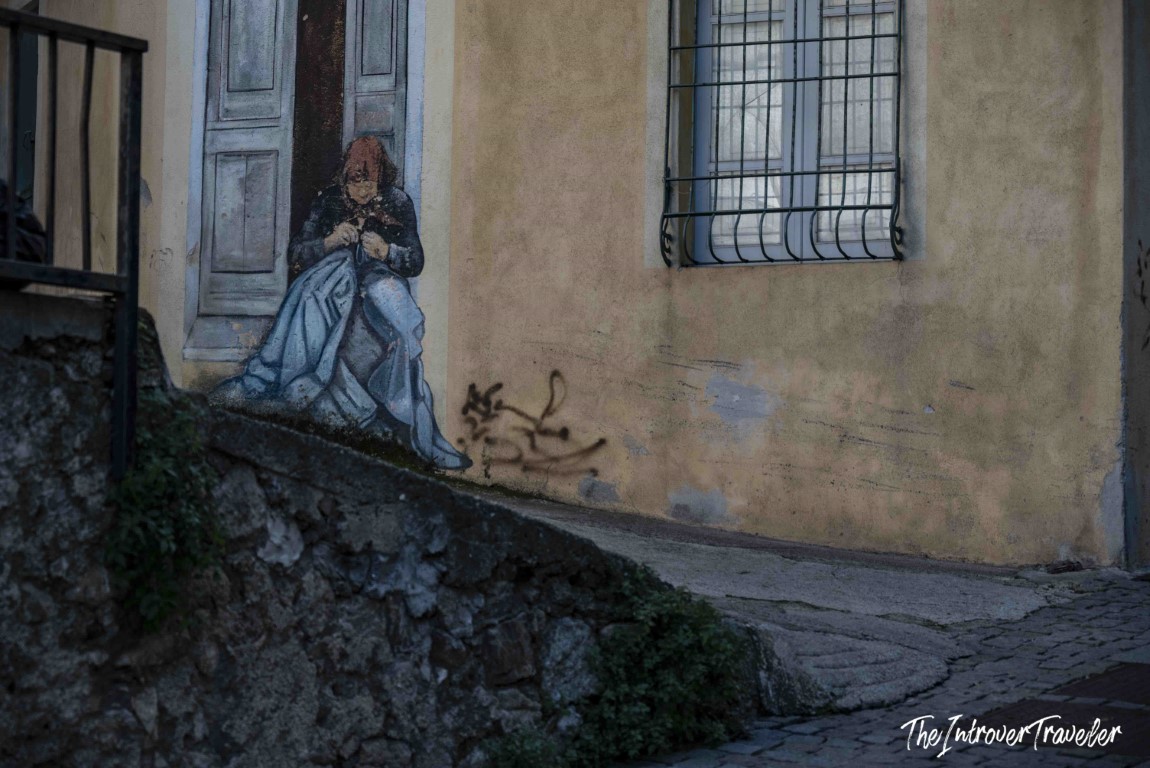 Street mural in Orgosolo, Sardinia, depicting a seated woman sewing beside a doorway