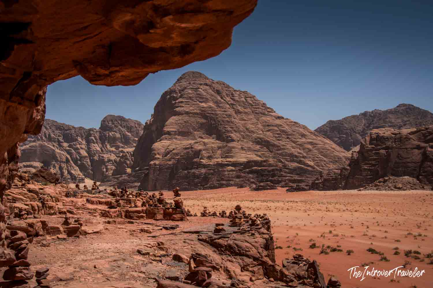 Wadi Rum desert landscape framed by a rock overhang, with stone cairns and red sand plains below layered sandstone mountains