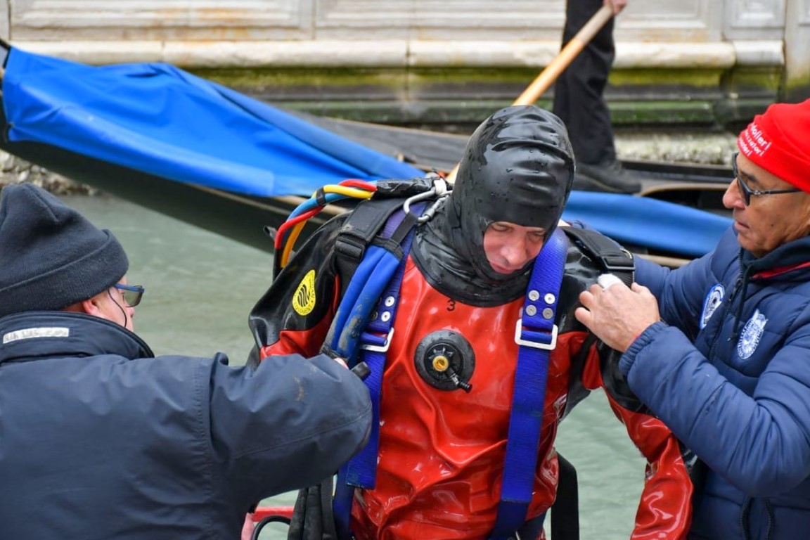 Diver in red dry suit being helped by two assistants beside a Venice canal gondola
