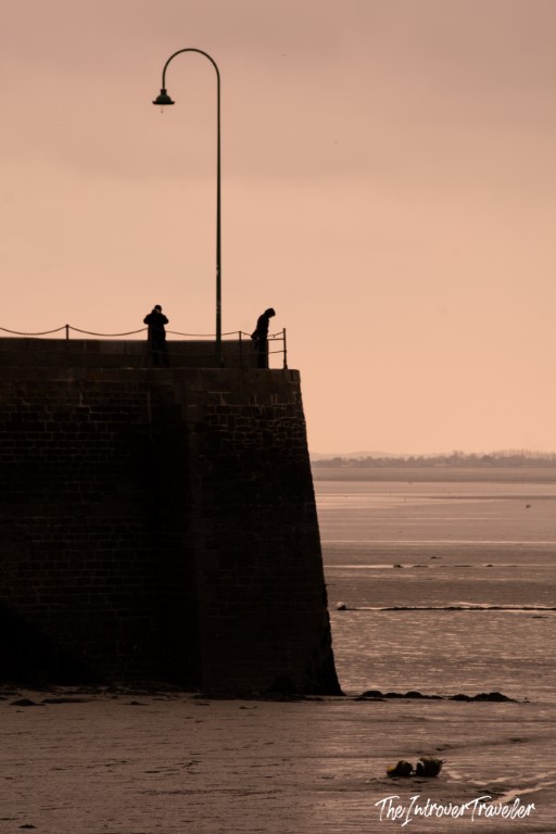 Two silhouettes on Cancale's stone harbour pier at dusk, with calm bay waters stretching behind