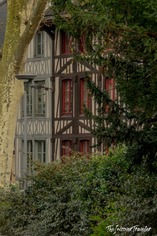 Half-timbered medieval building with red shutters peeking through lush greenery in Rouen, Normandy