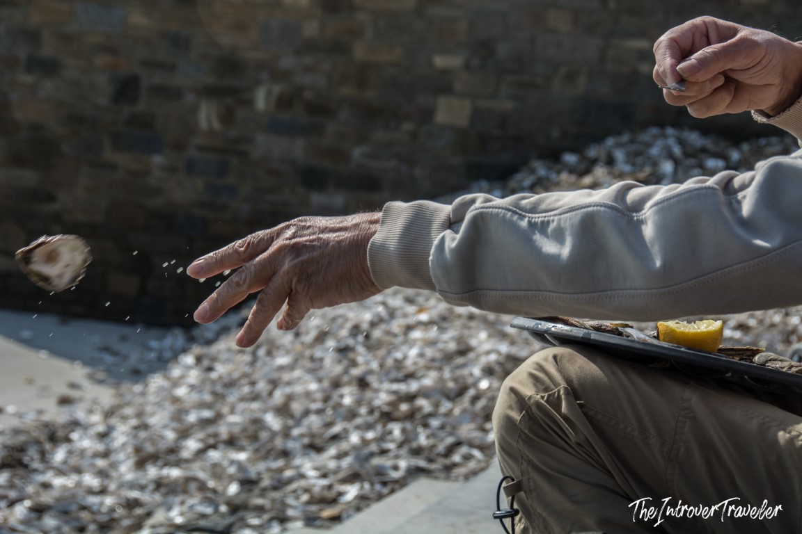 Cancale oyster shucker tossing a shell onto the seafront pile, with lemon and knife nearby