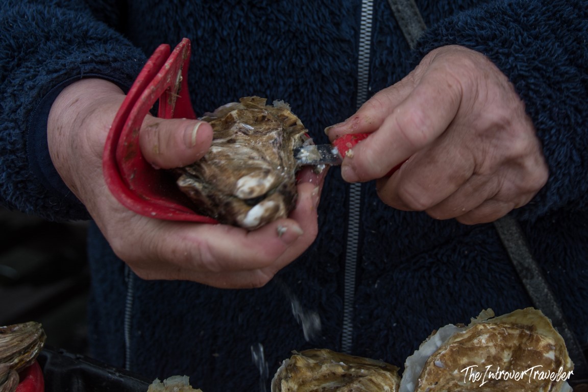 Vendor shucking a fresh Cancale oyster with a red-handled knife at the Brittany market
