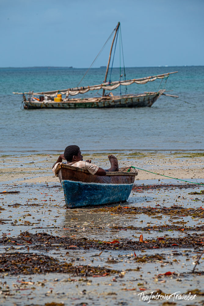 Boy resting in a small wooden boat at low tide on Mafia Island, Tanzania, with a traditional dhow anchored offshore