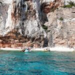 Turquoise water below white and orange limestone cliffs with a small beach and moored boats, Gulf of Orosei, Sardinia